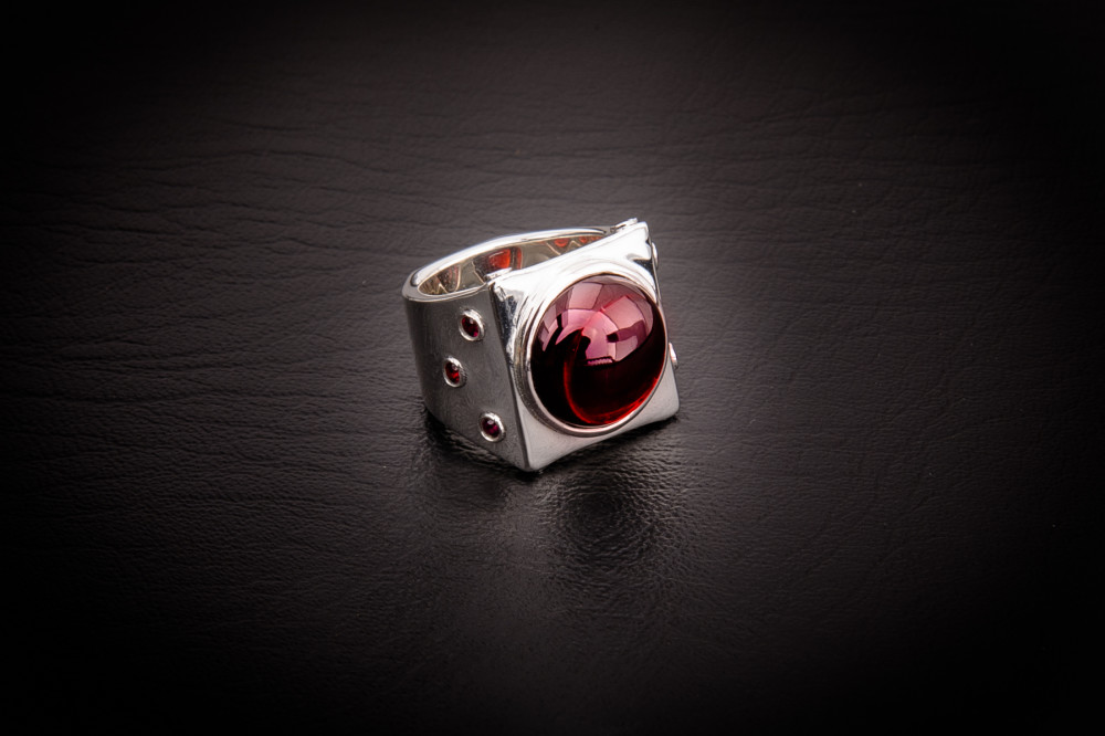 The Carson Ring is a contemporary ring from Leslie Herbert Jewelry. It features a large smooth garnet cabochon with smaller faceted garnets surrounding. A wide ring with modern elegant style. Red stones set in sterling silver. Fine jewelry from The Haviland Collection.