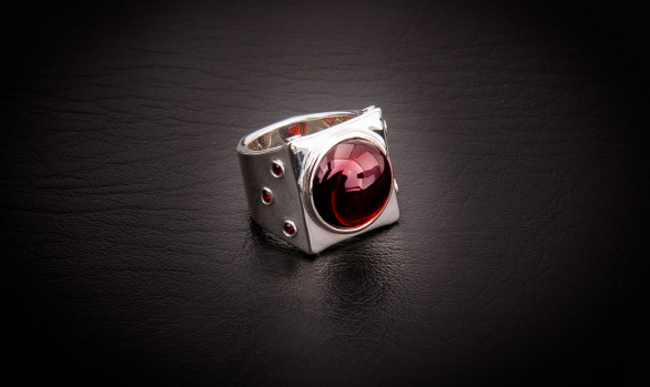 The Carson Ring is a contemporary ring from Leslie Herbert Jewelry. It features a large smooth garnet cabochon with smaller faceted garnets surrounding. A wide ring with modern elegant style. Red stones set in sterling silver. Fine jewelry from The Haviland Collection.